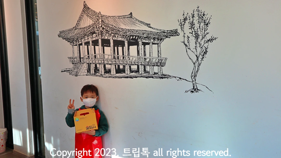 하얀 벽에 한옥과 나무가 검정색으로 그려져있고 그 아래 아이가 완성된 초코파이를 들고 있다.
