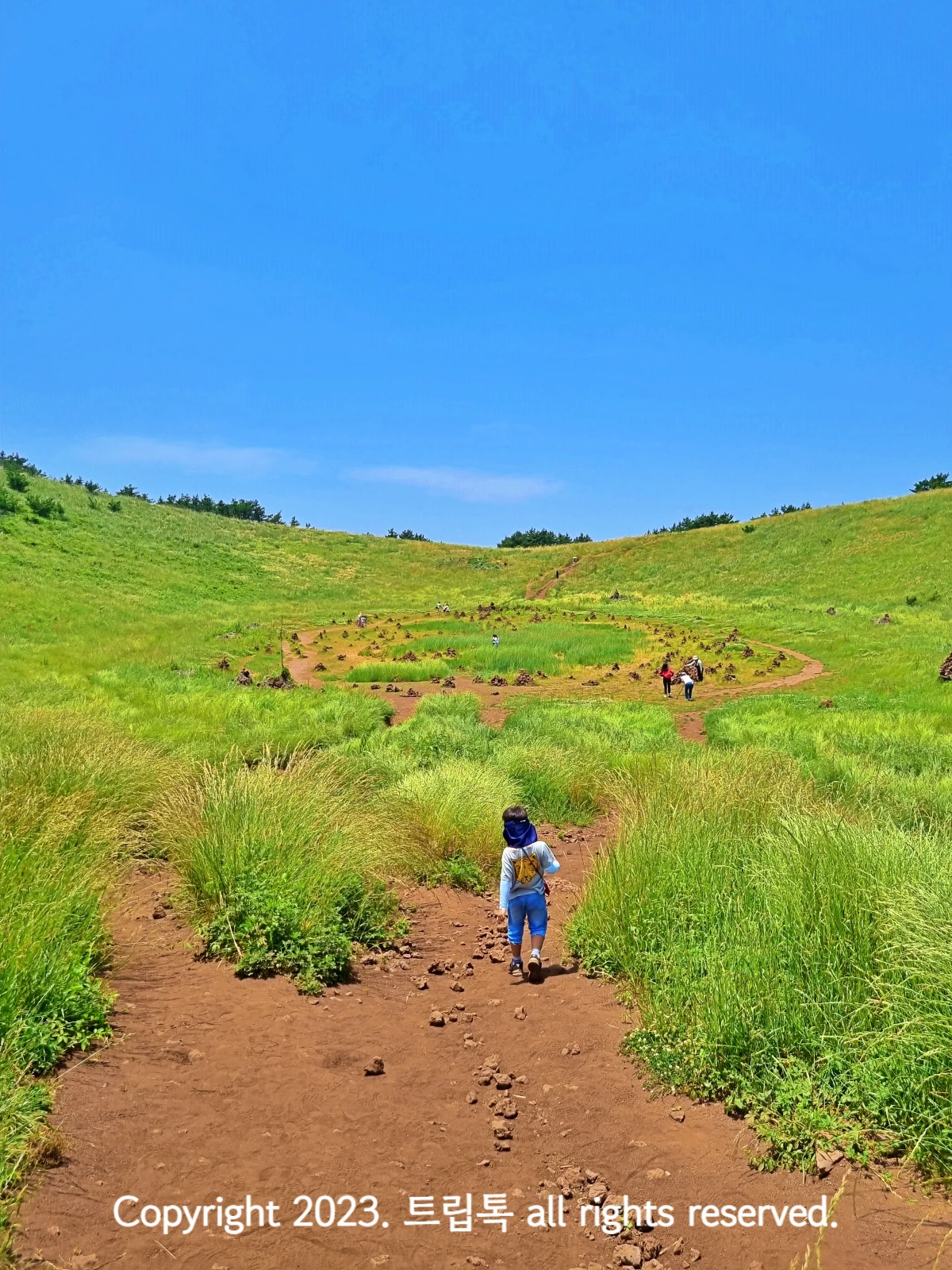 연두색 풀로 뒤덮인 산 아래 분화구를 흙길을 따라 내려가는 아이의 뒷모습