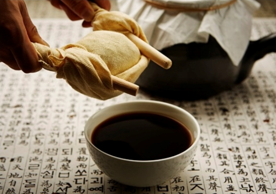 옛글자가 적혀있는 종이 위에 옹기약탕기를 놓아두고 그 밑에는 삼베 약보자기를 나무젓거락으로 짜고 있는 손과 그 밑에는 동그랗고 하얀 그릇에 검은 한약이 가득 담겨 있는 것을 찍은 사진