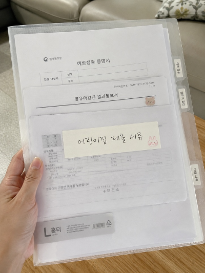 예방접종 증명서와 영유아검진 결과통보서가 인덱스 탭으로 분류되어 투명 L홀더 파일에 담겨 있고 겉면에 어린이집 제출 서류 메모가 붙어 있는 모습