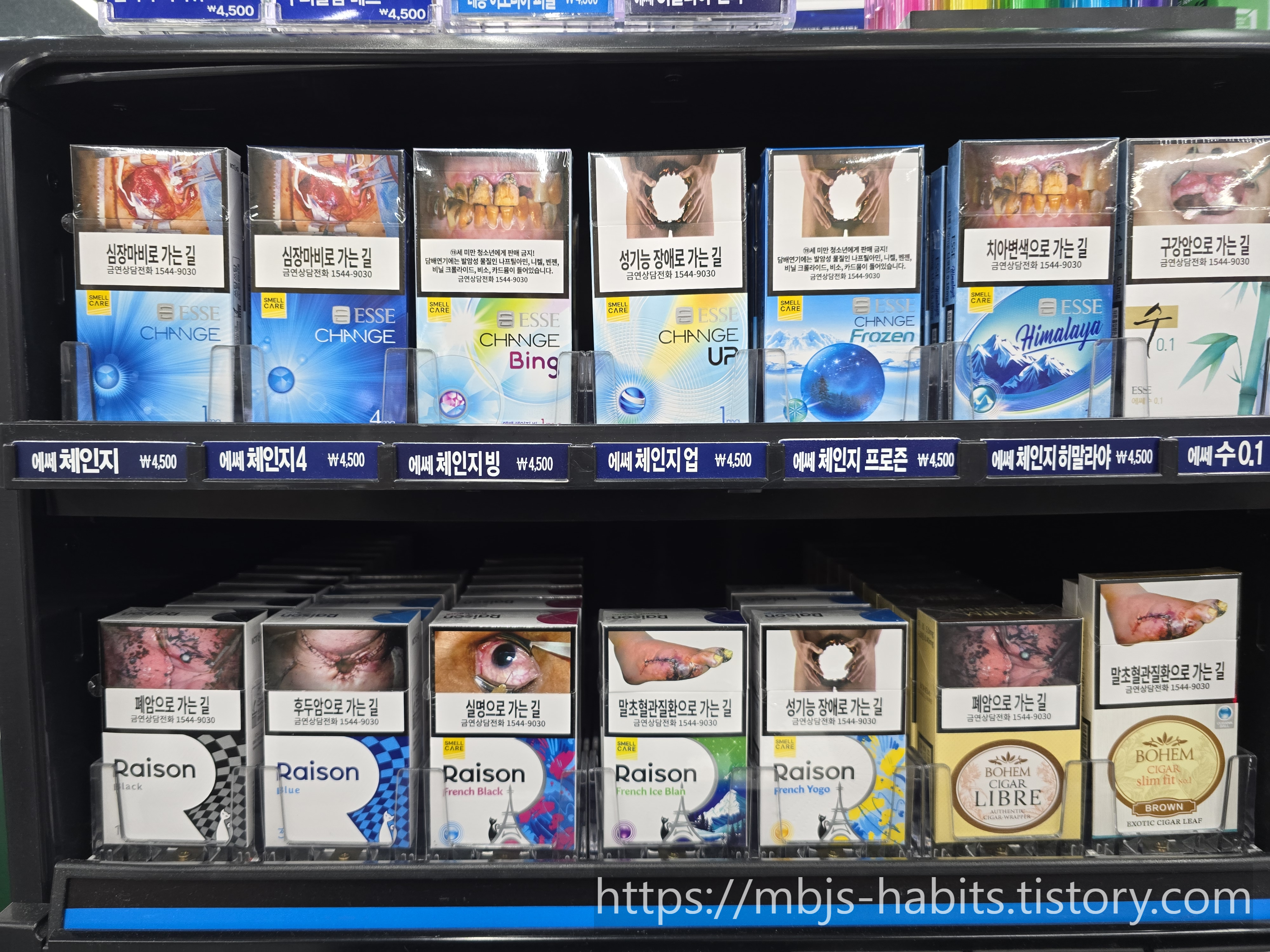 편의점 담배 진열대에 놓인 다양한 담배 제품과 경고 이미지 - 심장마비, 폐암, 성기능 장애, 실명, 구강암 등 흡연 관련 질환 경고 문구가 표기된 담배갑들