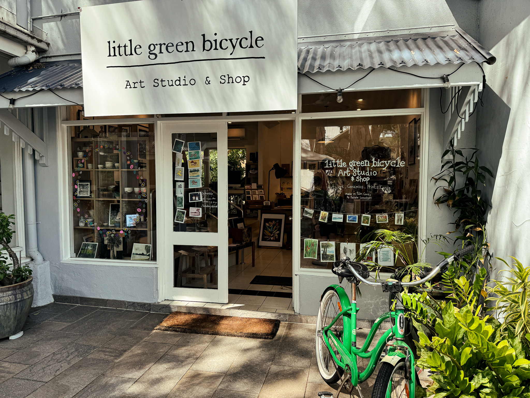"little green bicycle — Art Studio & Shop" 간판이 달린 팜코브 아트 스튜디오 & 숍 외관 — 흰 벽, 그림과 소품이 진열된 유리 쇼윈도, 입구 앞에 초록색 자전거가 세워진 모습
