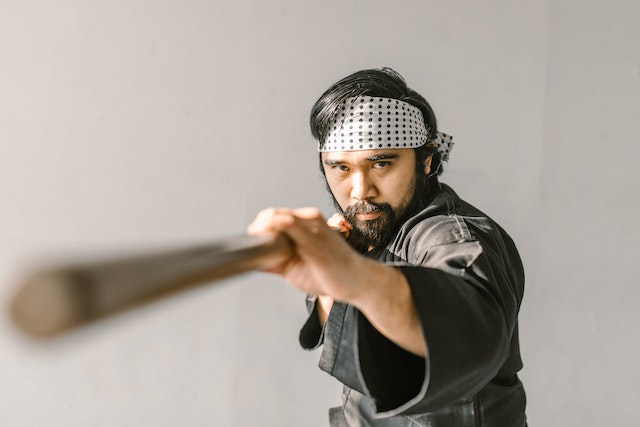 A man with a headband and long beard is pointing forward with a wooden pole for defense.
