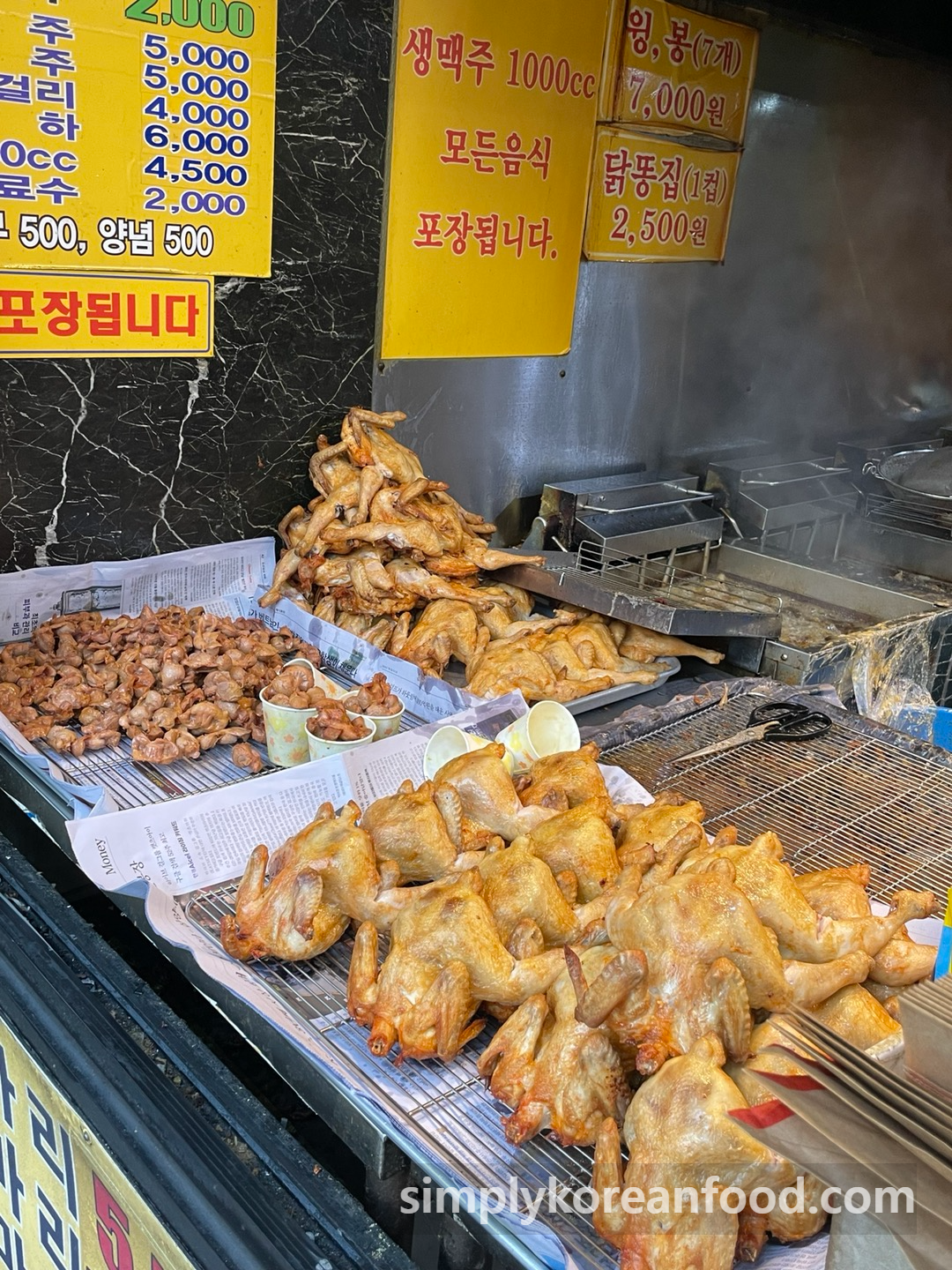 Rows of whole Korean fried chickens cooling on metal trays outside Korea Tongdak in Dongmyo, with chicken gizzards and wings for sale nearby.