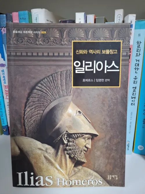 "호메로스의 『일리아스』 줄거리와 배경을 시각화한 예술적 이미지. 노을 지는 트로이 해변에 놓인 아킬레우스의 투구와 방패, 그리고 그 곁에서 빛을 머금은 채 기록을 기다리는 고대 양피지 두루마리를 통해 운명에 맞선 영웅들의 서사와 기록을 통한 지적 자립의 메시지를 상징함."