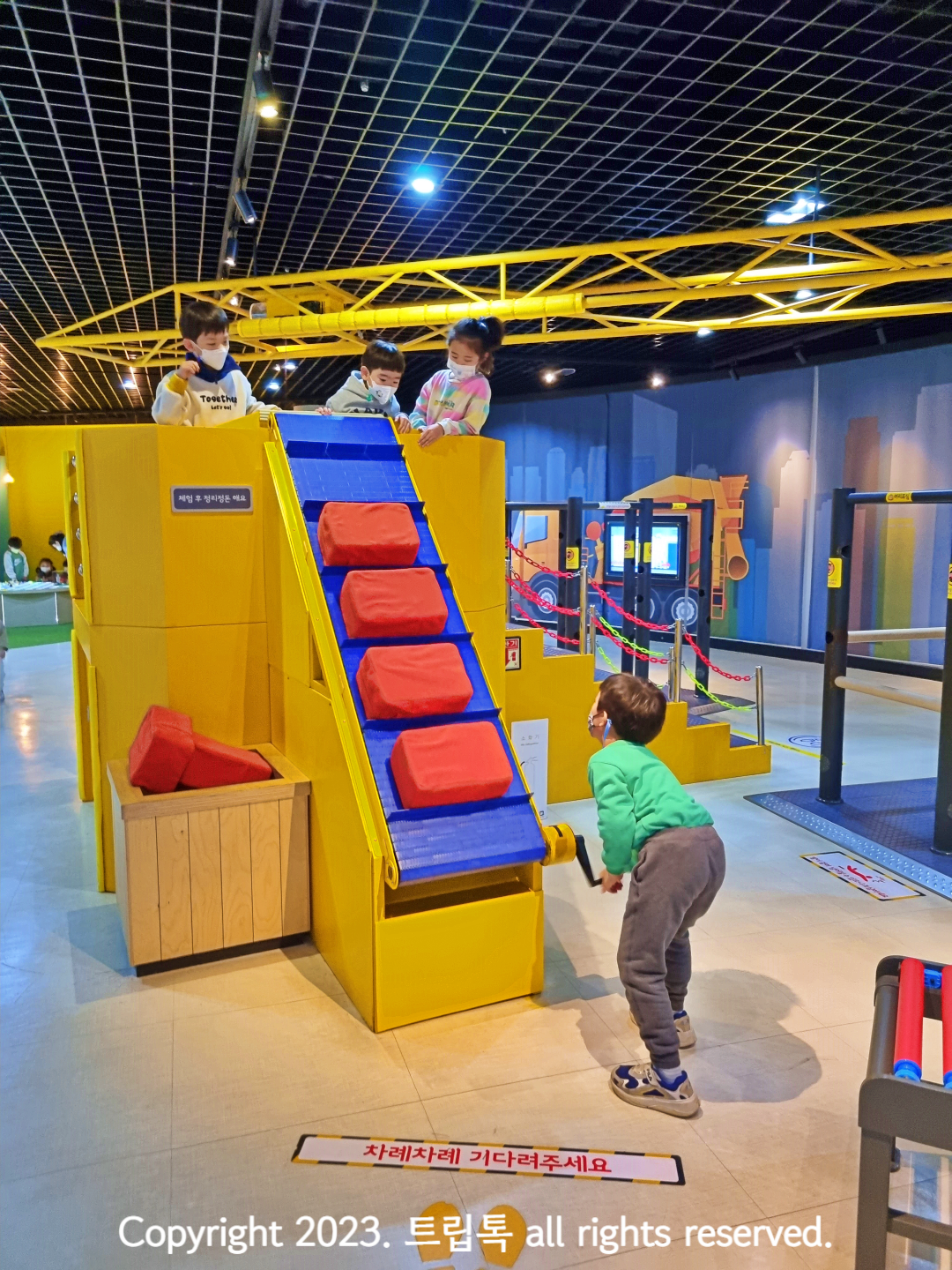 아래에서 아이가 빨간 쿠션을 놓고 손잡이를 돌리면 레일을따라 움직이는 쿠션을 위의 아이들이 기다리고 있다.