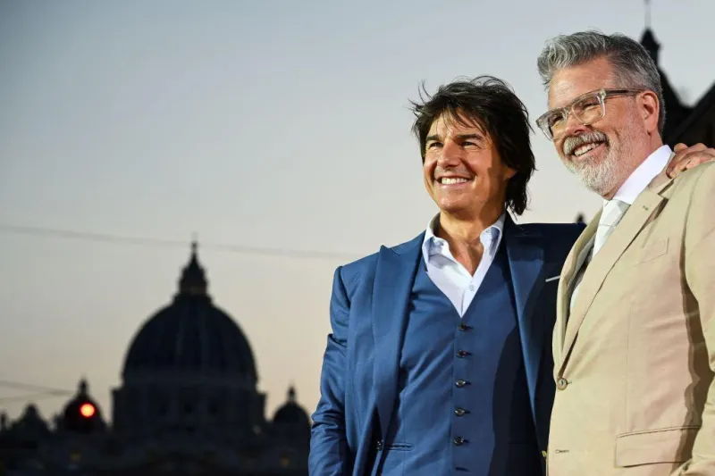 야외를 배경으로 파랑 정장을 입은 톰크루즈와 베이지색 정장을 입은 크리스토퍼 매쿼리 감독이 어깨동무를 하면서 웃고 있는 모습