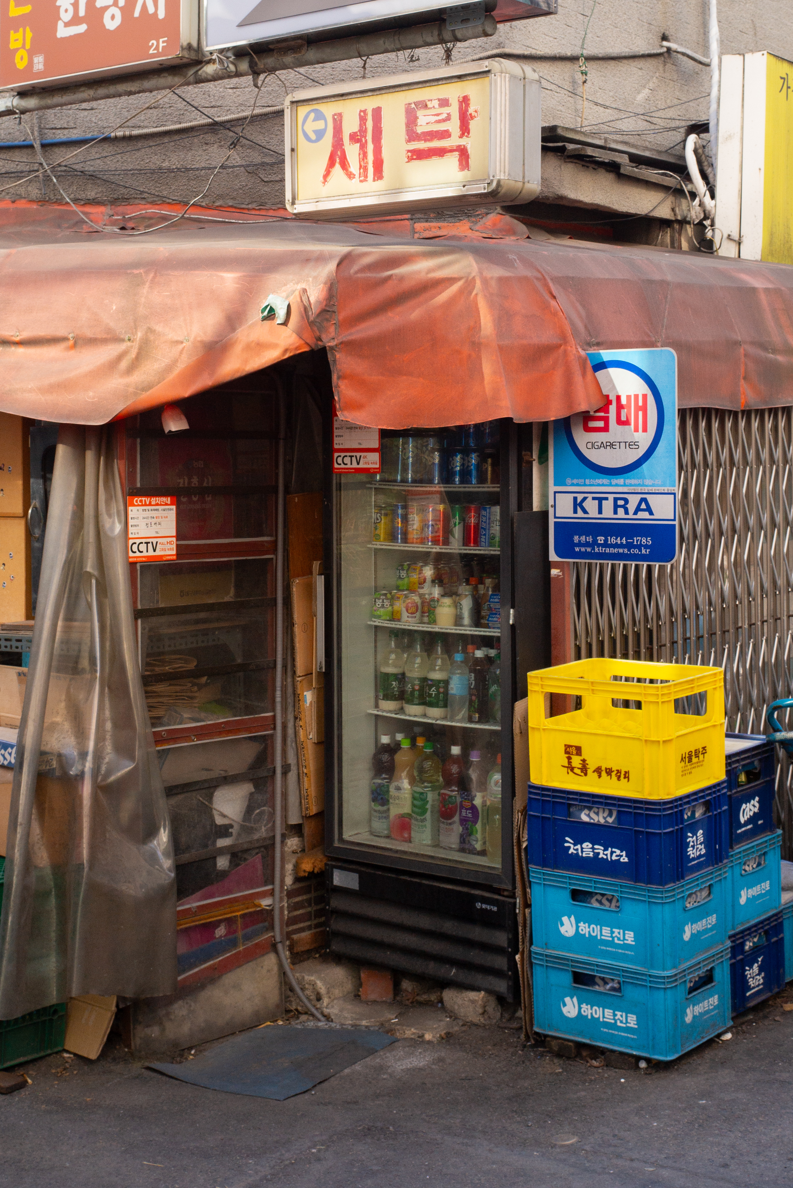 '세탁'과 '담배' 간판이 있는 노포 입구에 쌓인 파란색과 노란색 플라스틱 상자들, 그리고 음료 냉장고가 만드는 일상의 질감