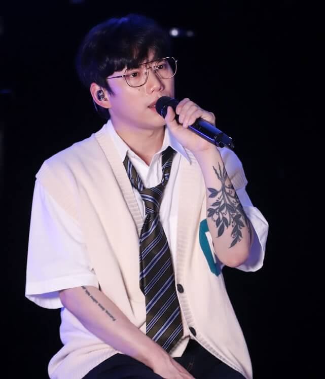 권정열이-하얀-셔츠에-박시한-조끼를-입고-왼손에-마이크를-들고-노래를-부르는-사진으로-그의-팔에는-꽃무늬-타투가-있다