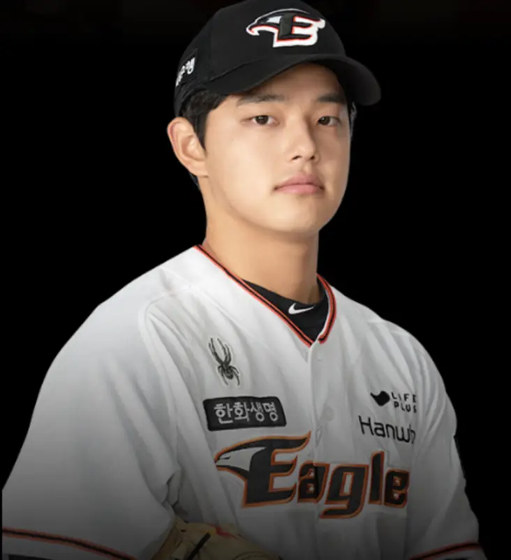 검은배경 검은 야구모자 가운데 흰테두리 검은마크 E 흰색 한화이글스 유니폼을 입고 옆으로 선채 정면을 바라보는 문동주