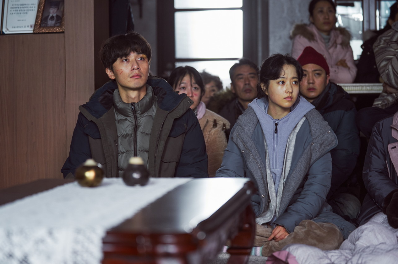 민성과 명화가 나란히 앉아 굳은 표정으로 앞을 바라보는 장면, 뒤로 다른 주민들이 보인다