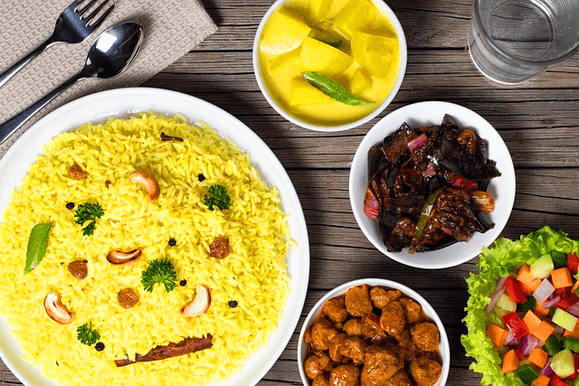 하얀색의 둥그런 접시에 노란색 강황밥이 가득하고, 옆 접시들에는 야채샐러드, 감자반찬, 가지볶음 등이 있다.