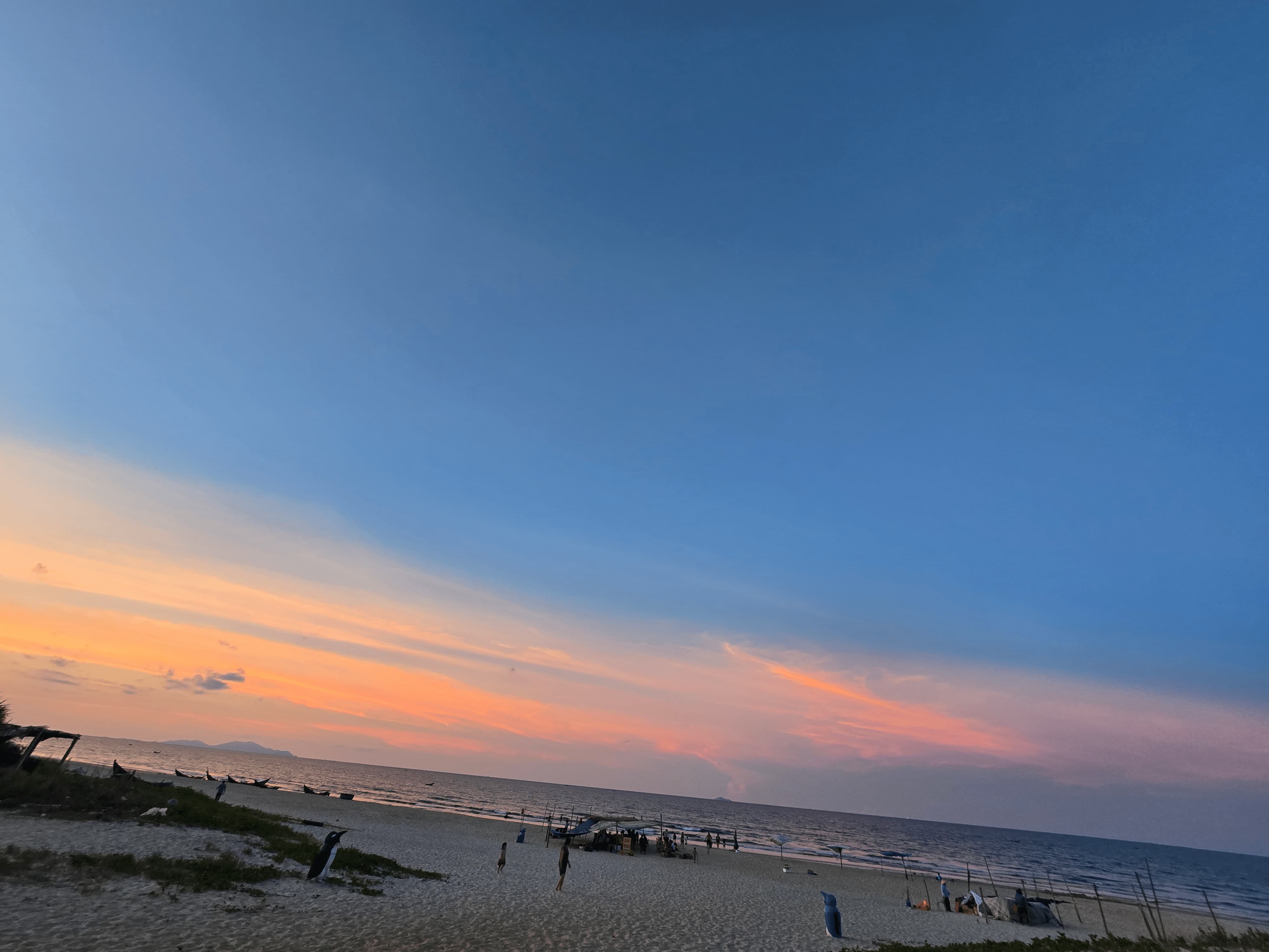 베트남 다낭 땀끼 해변의 일몰 풍경. 푸른 하늘과 붉은 노을빛이 어우러져 수평선 위로 물들고, 모래사장 위에는 사람들이 저녁 시간을 즐기고 있다. 바다 위에는 작은 배들이 보이며 평화로운 분위기를 전한다.