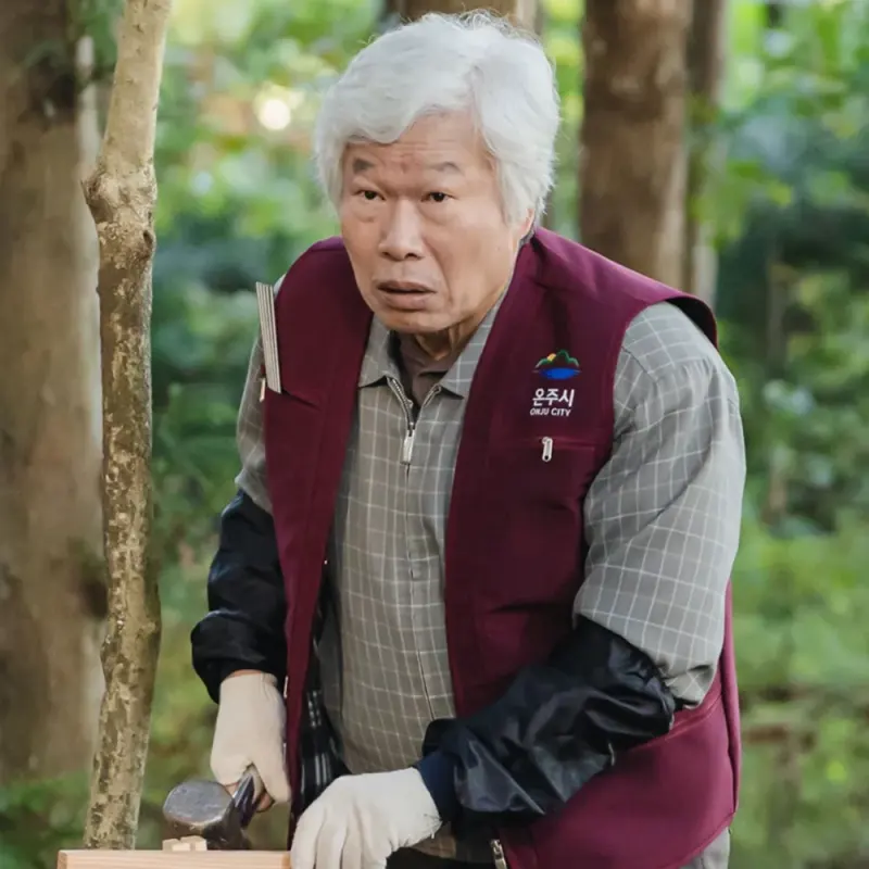나무가 우거진 곳을 배경으로 작업복을 입고 망치질을 하고 있는 백발의 드라마 이 연애는 불가항력 오삼식 캐릭터