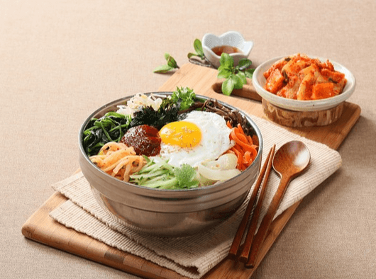 둥근 은그릇에 비빔밥이 가득하다. 위에는 발간 고추장, 노란 달걀프라이, 각종 야채나물들이 있고 주변엔 나무 젓가락과 숟가락, 깍두기가 있다.