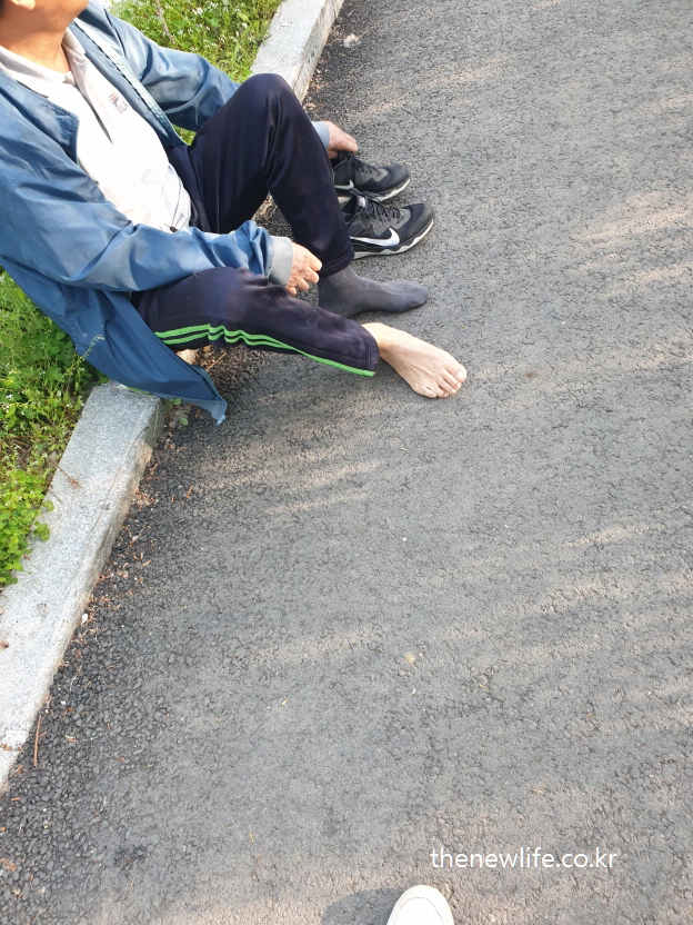 An elderly man sitting on the edge of a curb, taking off his shoes and socks before barefoot walking.-맨발 걷기를 시작하기 위해 신발과 양말을 벗고 있는 어르신의 모습. 인도 가장자리에 앉아 있는 장면