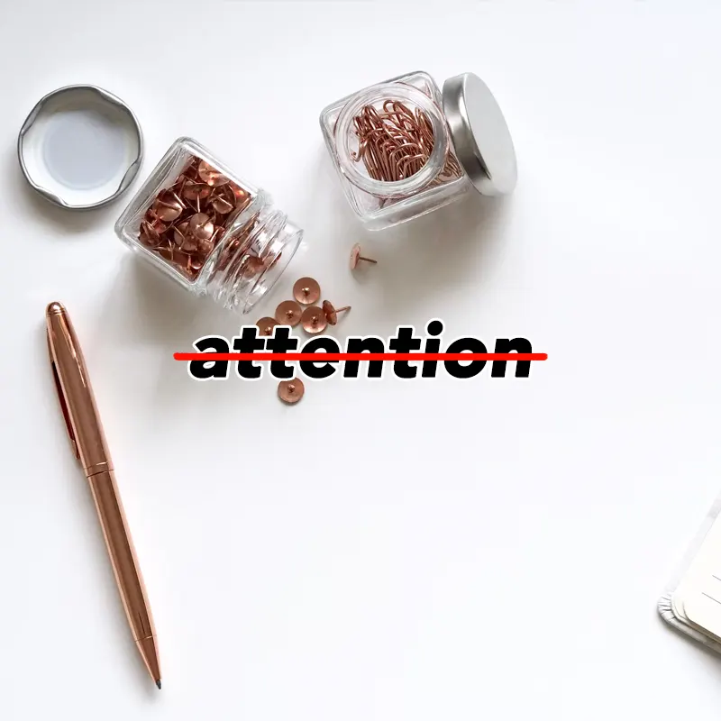 사무용품인 압정과 만년펜이 흰 책상 위에 있고, attention이라는 글자에 취소선이 그어져 있다.