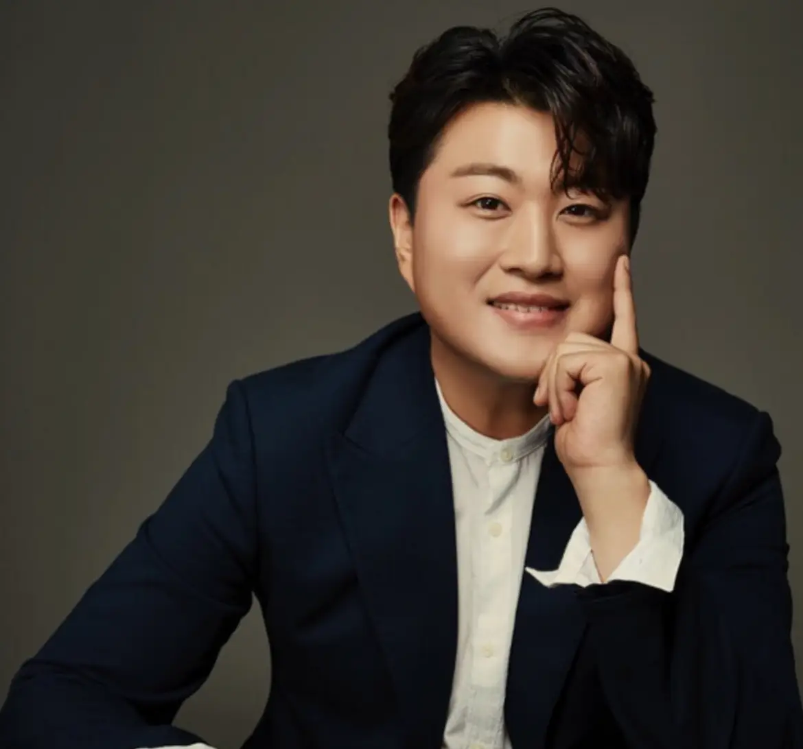 초록배경 검은 수트 흰 셔츠를 입고 왼손으로 턱을 괴고 미소짓는 모습