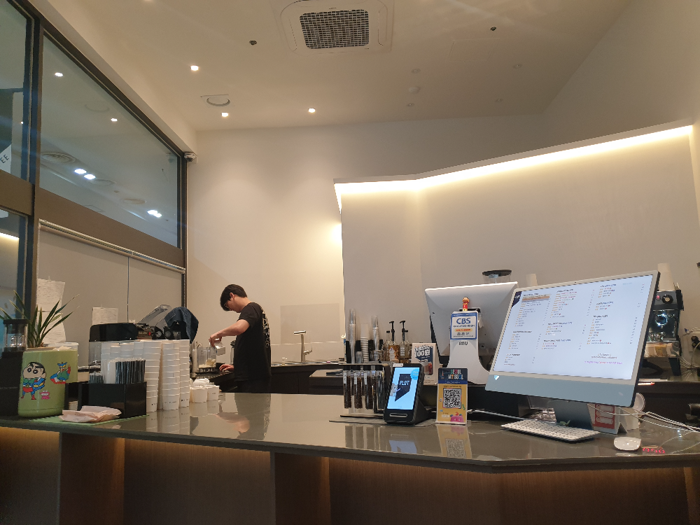 흰 천장과 벽면 그리고 중앙 테이블 뒤쪽 작업대에서 바리스타가 음료를 추출하고 있다.