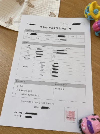 거실 테이블 위에 놓인 영유아 건강검진 결과통보서 종이 서류와 주변에 놓인 아기 장난감 자동차 및 딸랑이 소품 사진