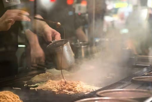 철판위에 음식이 있고 그 위로 소스를 뿌리고 있다 철판 주위에서 연기가 나고 있다.