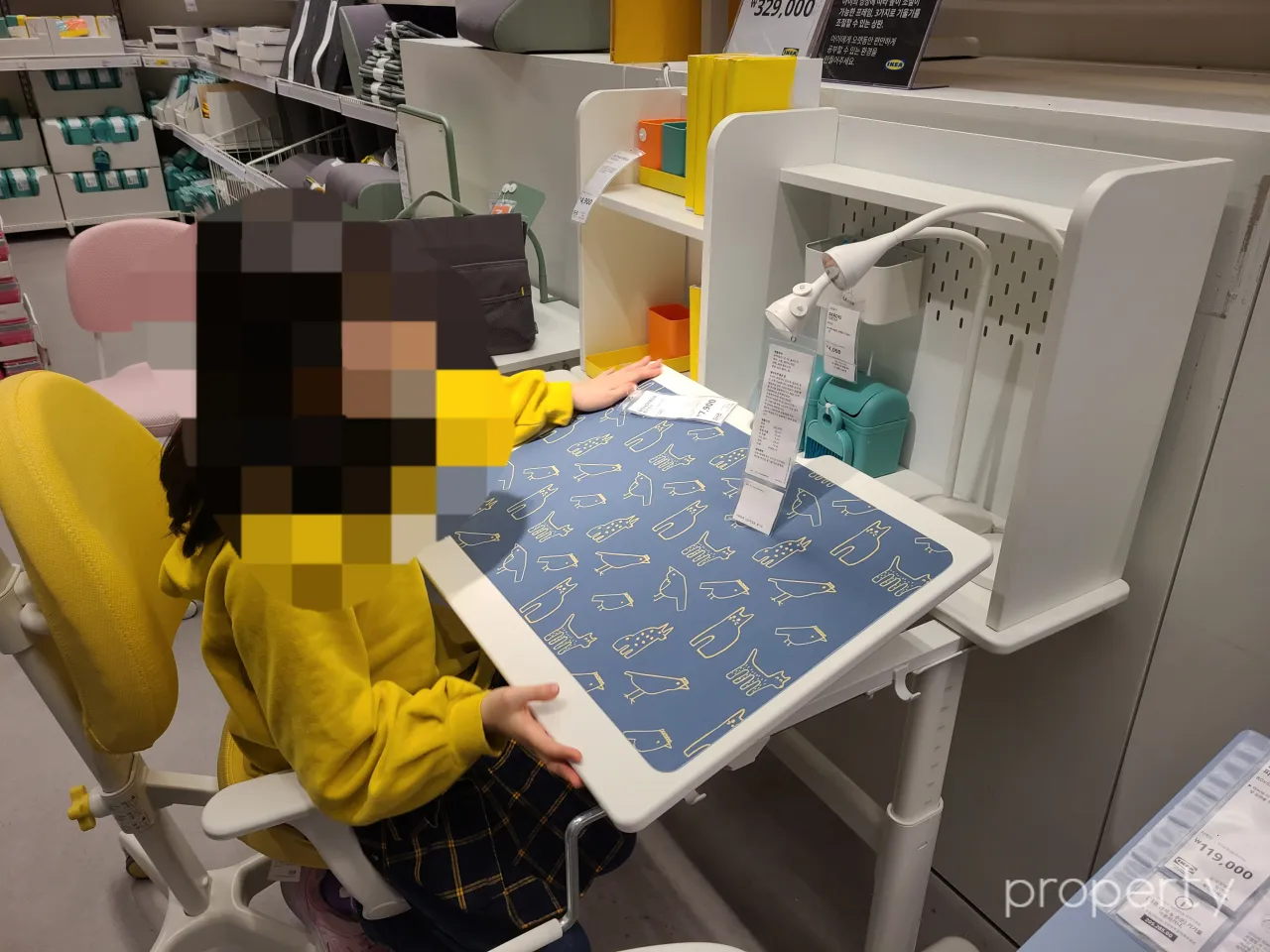 이케아매장안-책상작업대를세워놓고-노란색옷입은여자아이가앉아있다-책상위에민트색연필깍기-주황노랑연필꽂이-흰색스텐드가있는