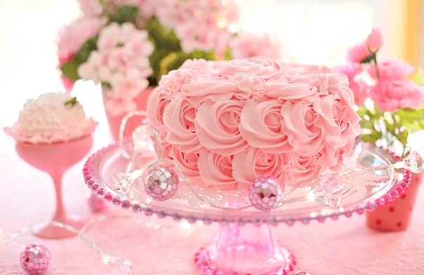 분홍색 생크림으로 장식한 예쁜 생일케이크가 있고 그 옆에는 분홍 꽃들이 가득하다.