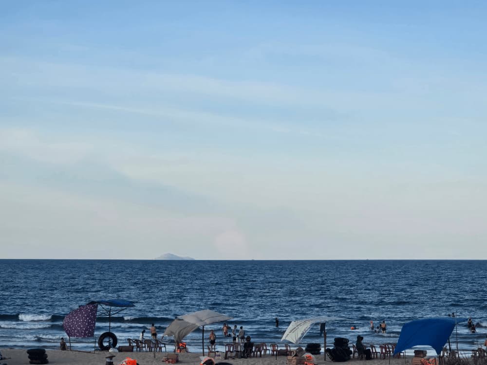 베트남 다낭 땀끼 해변 풍경. 파라솔과 의자들이 늘어선 모래사장에서 사람들이 휴식을 즐기고, 바다에서는 현지인들이 물놀이를 하고 있다. 멀리 수평선에는 작은 섬이 보이며, 여름 바다의 여유로운 분위기를 담고 있다.