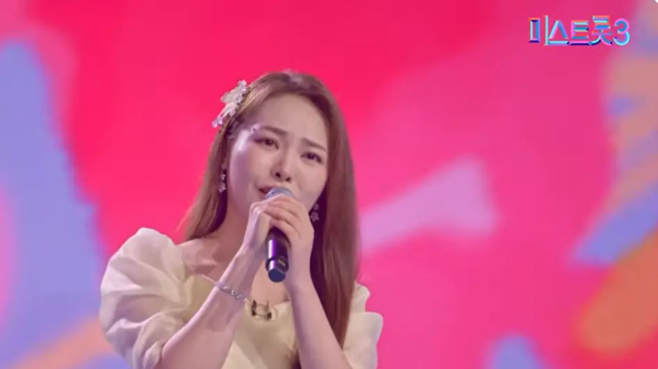미스트롯3- 빨간배경 노란 드레스를 입고 긴머리를 늘어뜨린채 두손으로 마이크를 잡고 노래하는 미스김