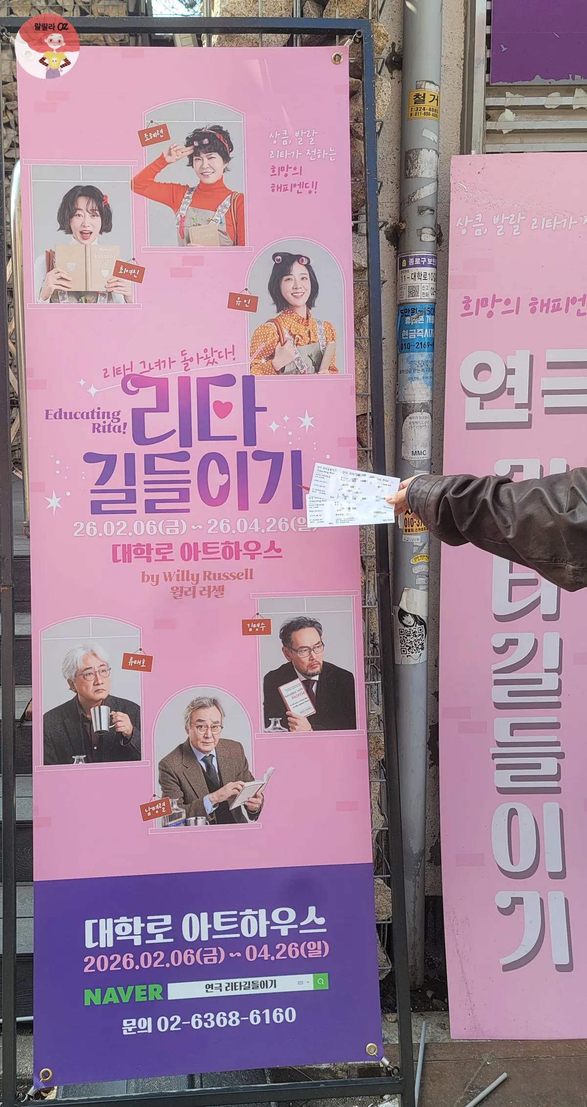 리타길들이기 배너 광고를 배경으로 손에 든 티켓 두장,배너에는 남녀주인공들의 사진이 있다.