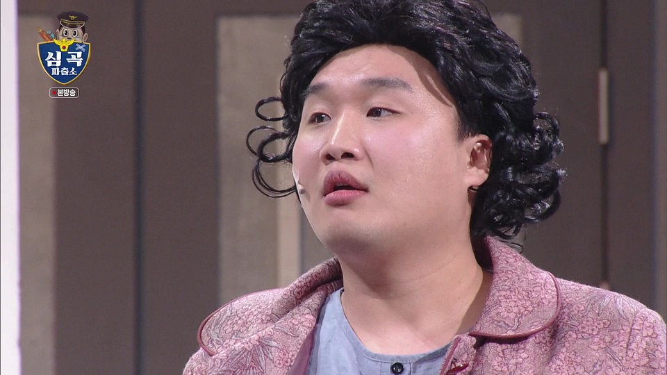 심곡 파출소 표시 아래 곱슬머리 가발과 분홍색 겉옷을 착용한 출연자가 보이는 방송 화면