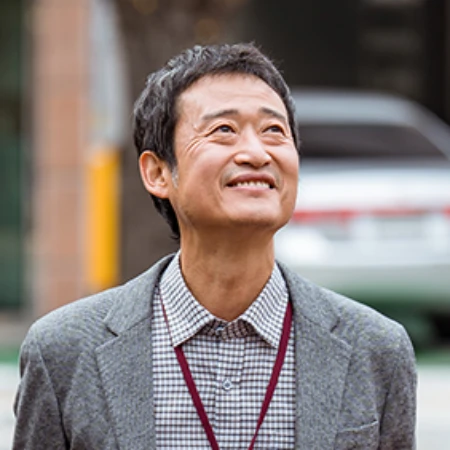 어느 실외를 배경으로 회색 자켓을 입고 새치가 많이 난 오늘도 사랑스럽개의 교감 캐릭터