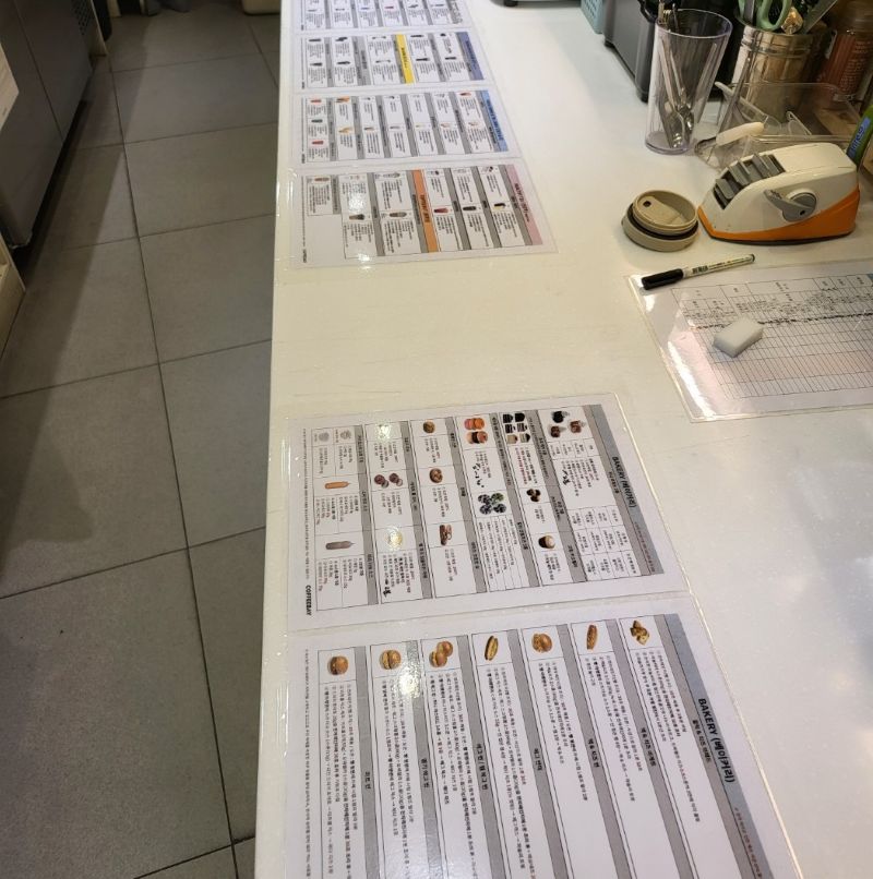 카페 작업대(조리대) 위에 여러 장의 코팅된 레시피 카드가 일렬로 길게 부착되어 있는 모습. 조리 동선에 맞춰 재료 계량과 동시에 시선을 내리면 바로 수치를 확인할 수 있도록 배치되어 있어, '찾는 시간'을 없애고 오염을 차단하는 정갈한 시각화 시스템의 실체를 보여줌.