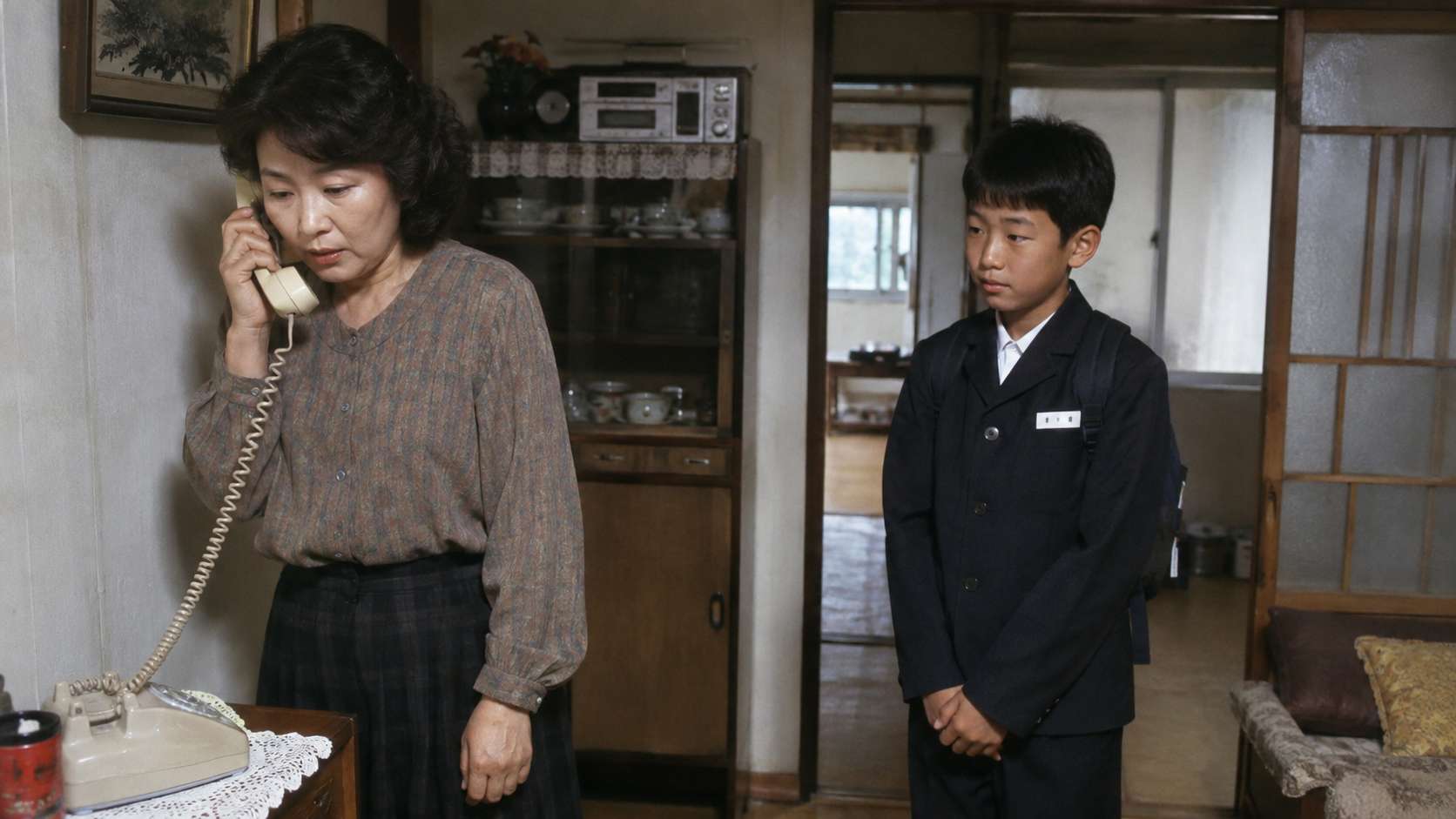 집 안에서 어머니가 집 전화를 받고 있고, 교복 차림의 아이가 옆에서 차례를 기다리는 모습