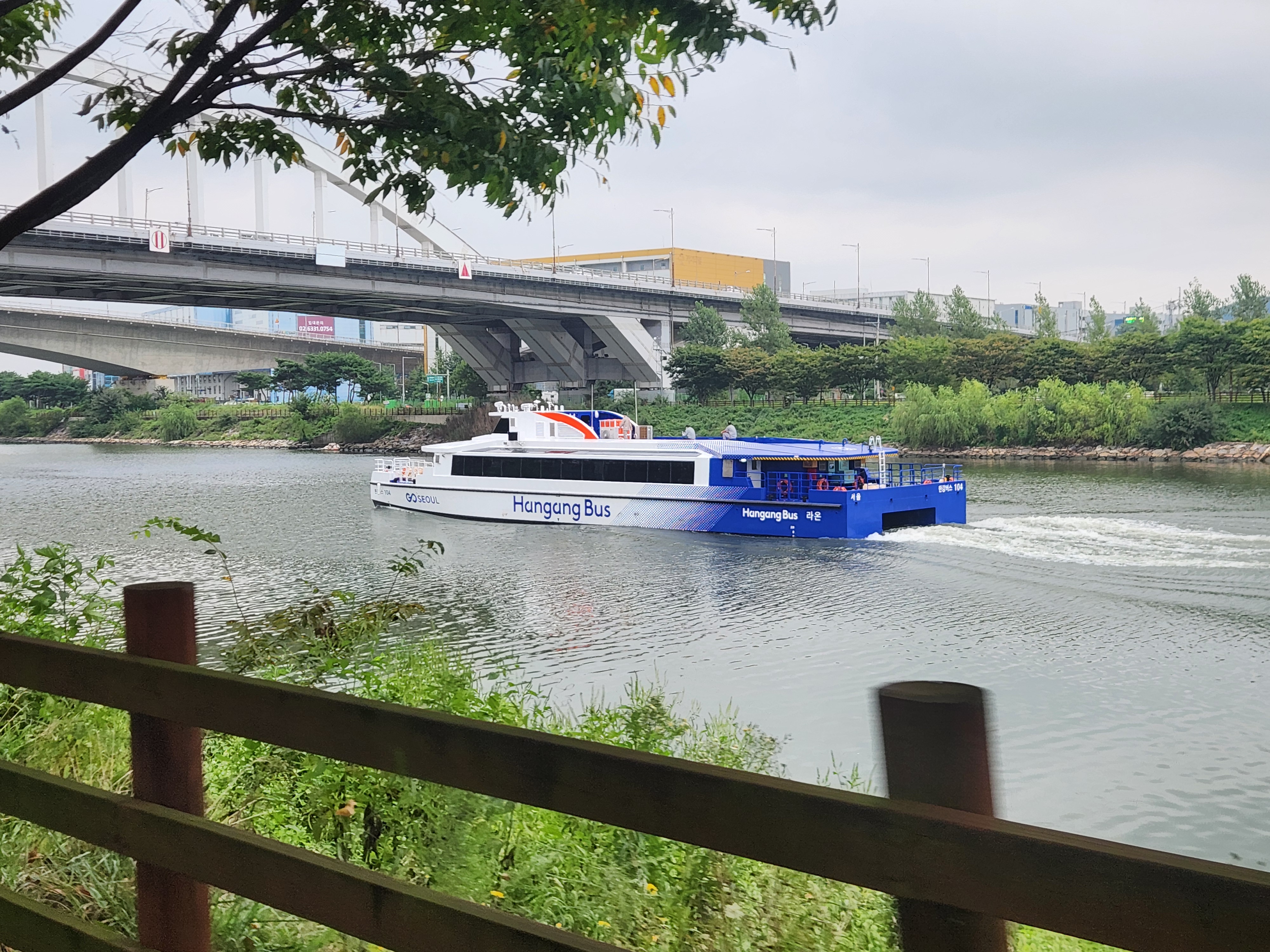 서울 한강 위를 운항 중인 파란색&middot;흰색 한강버스(Hangang Bus), 교각 아래를 지나며 물살을 가르는 모습