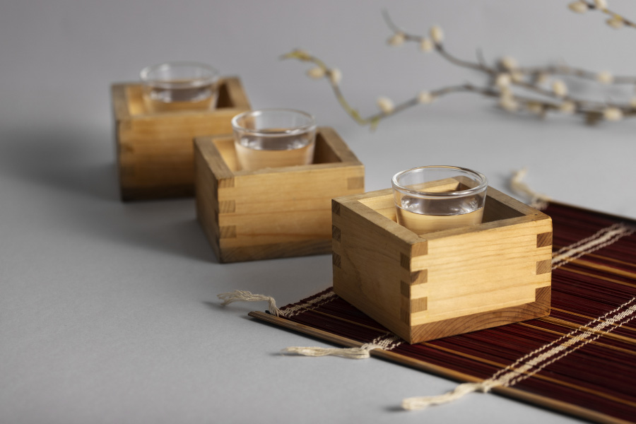 흰색 바닥 위에 투명한 청주를 가득 담은 투명한 잔을 담은 작은 나무 상자 3개와 대나무로 만든 깔판, 매화나무를 같이 놓아두고 찍은 사진