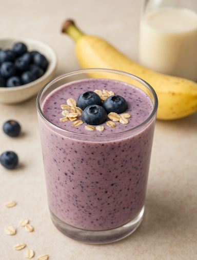 투명한 유리컵에 블루베리 귀리 스무디가 담겨 있는 모습. 토핑으로 블루베리와 귀리가 있다. 뒷배경으로는 작은볼에 담긴 블루베리와 바나나와 병에 담긴 아몬드 우유가 있다.