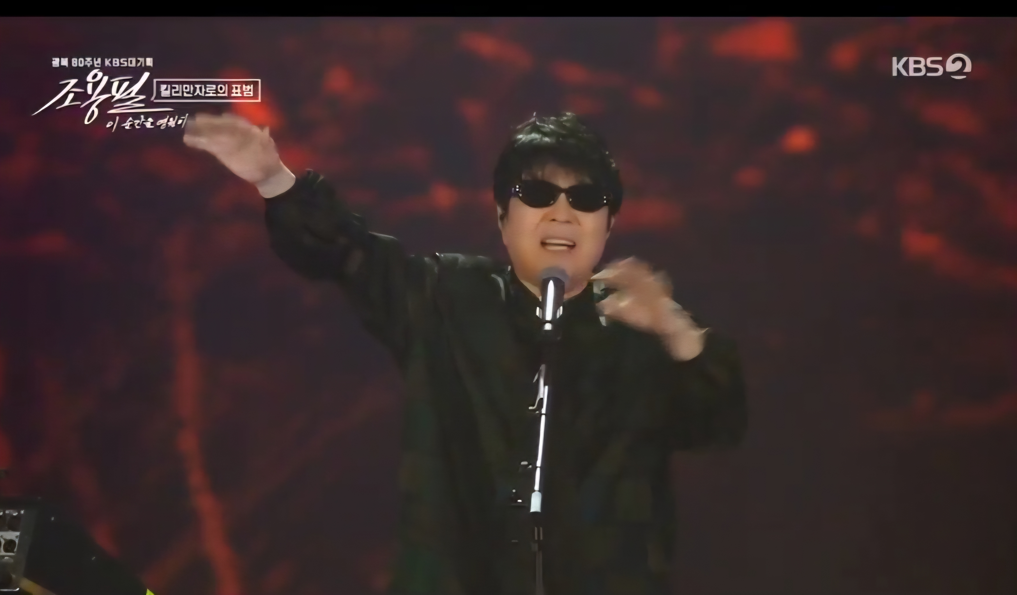 마이크를 잡고 손을 들어 올리며 노래하는 조용필의 모습