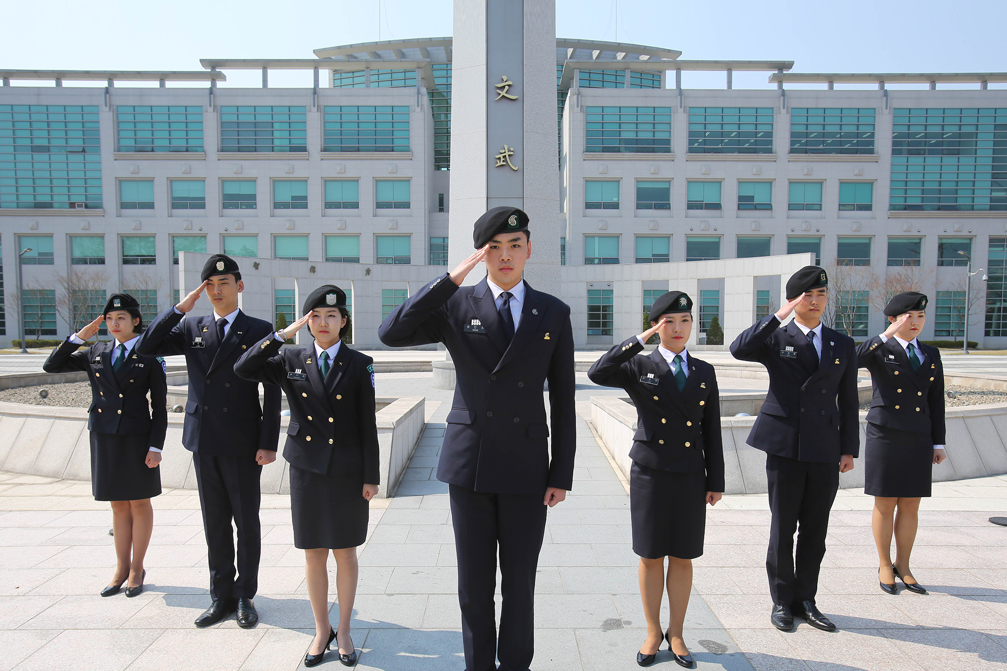 ROTC 사관후보생 남녀 7명이 모두 단복과 단모를 착용하고 오른손으로 경례를 하고 있는 사진, 뒤에는 괴산 육군중앙군사학교 정문이 보인다.