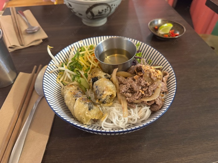 A freshly prepared bowl of Vietnamese Bo bun topped with crispy nems and savory beef on a wooden table ❘ Un bol de Bò bún vietnamien fraîchement préparé garni de nems croustillants et de bœuf savoureux sur une table en bois