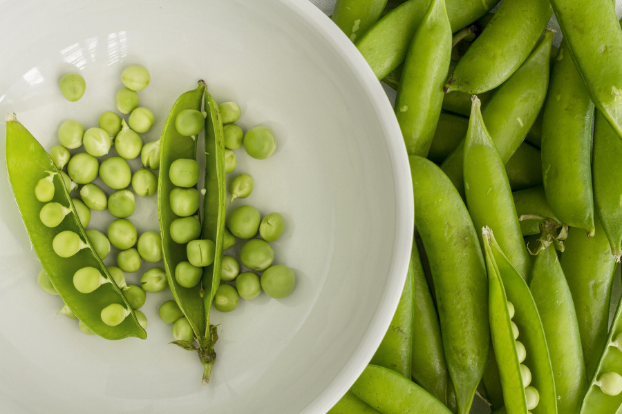 바닥에 완두콩 콩깍지를 깔아 놓고 그 위에 희고 동그란 그릇에 완두콩 콩까지를 까서 속에 완두콩이 보이게 2개를 놓아두고 그 주변에 완두콩을 여러 개 놓아두고 찍은 사진