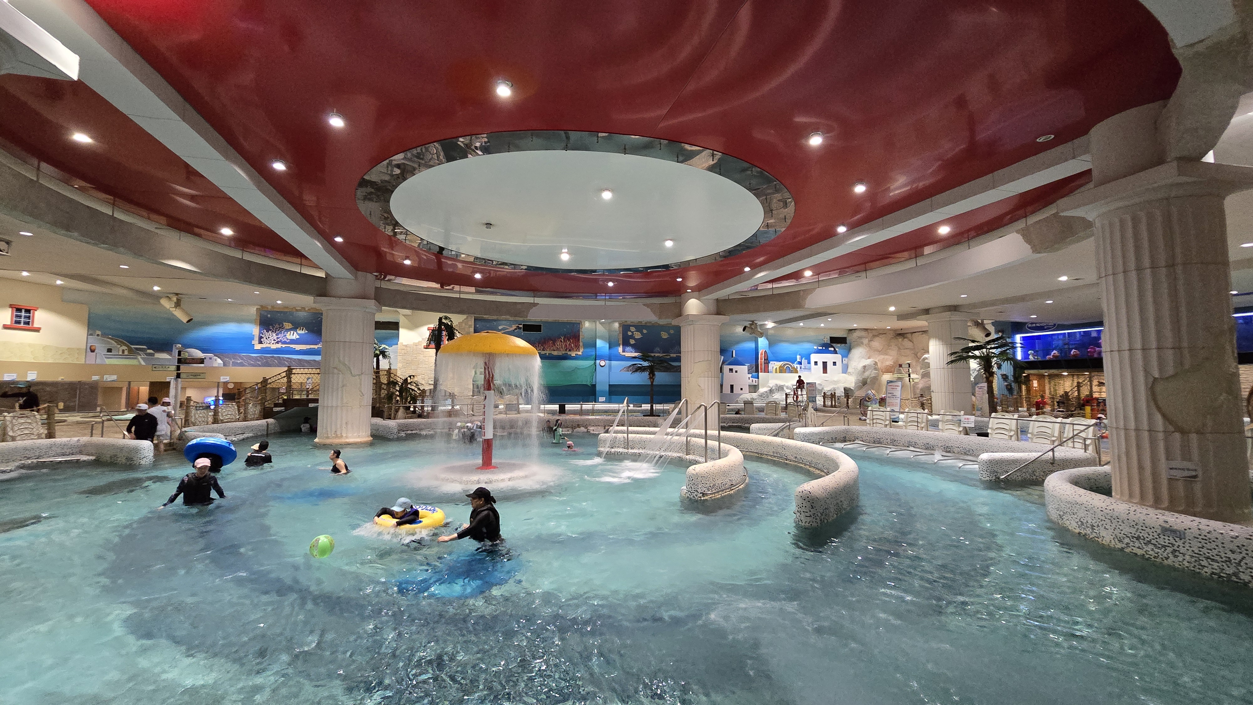 영등포 씨랄라 워터파크 내부 메인풀 전경. 산토리니 벽화와 분수형 물놀이 시설이 어우러진 실내 유수풀 공간으로, 가족 단위 이용객들이 튜브를 타고 물놀이를 즐기고 있다.