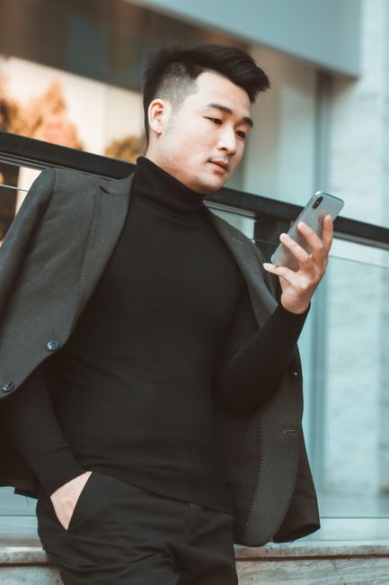 검은색 터틀넥과 블레이저를 입은 패셔너블한 남자가 야외에서 전화기를 확인하고 있습니다.