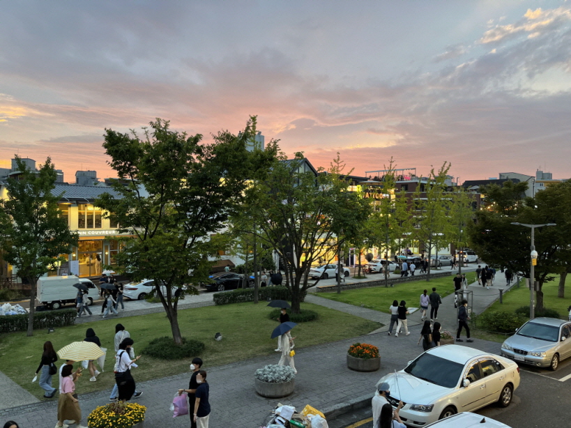 해질 무렵 분홍빛과 보랏빛이 어우러진 하늘 아래, 도심 속 공원과 상점가에 많은 사람들이 걷고 있는 풍경. 나무와 화단 사이를 걷는 사람들, 도로를 지나는 차량, 저녁 빛에 물든 평화롭고 활기찬 거리의 모습이 담겨 있다.