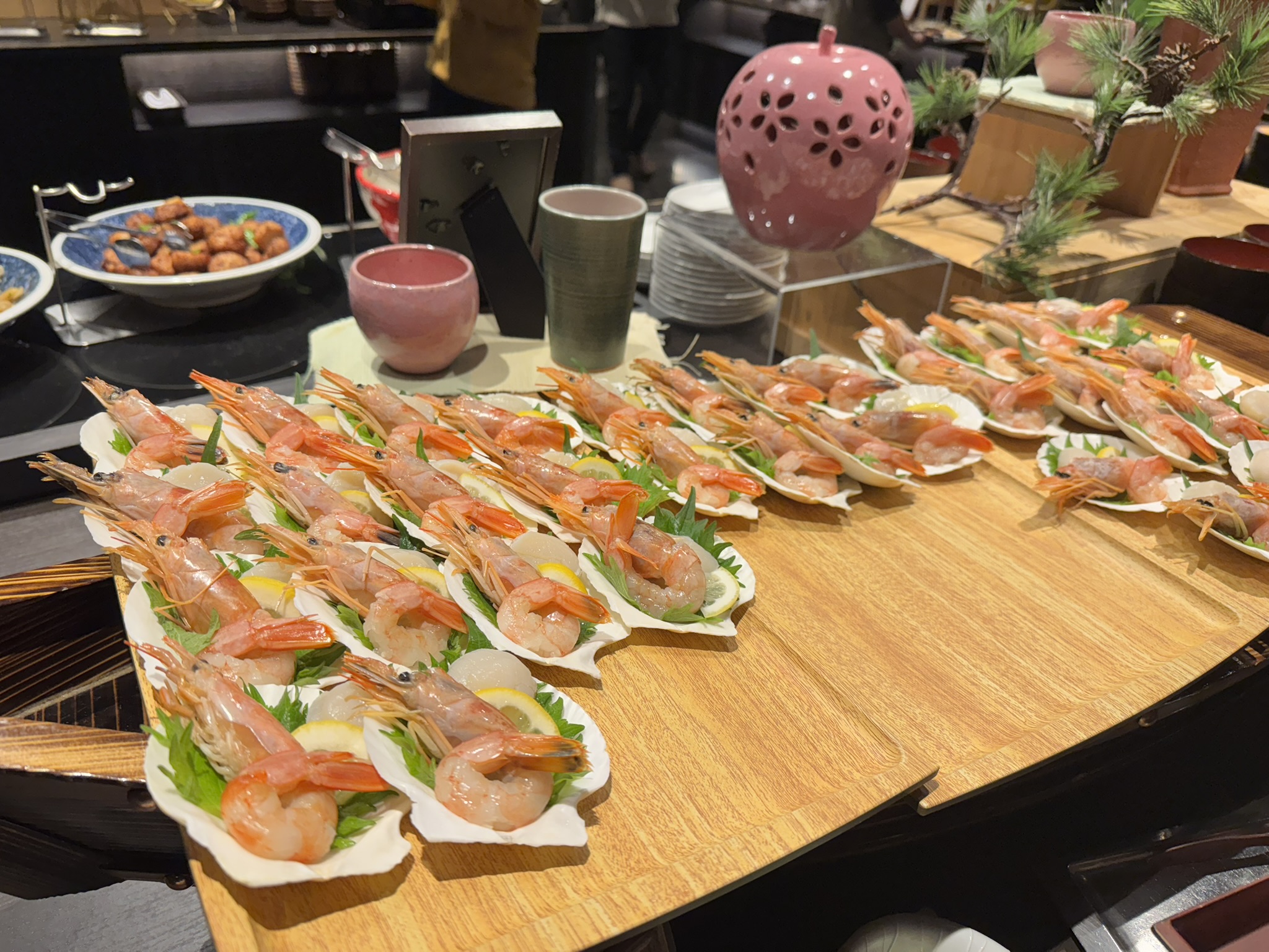 가리비 껍데기 위에 신선한 생새우와 레몬 슬라이스, 시소 잎이 정갈하게 세팅된 해산물 전채 요리들이 나무 선반 위에 줄지어 있는 모습.