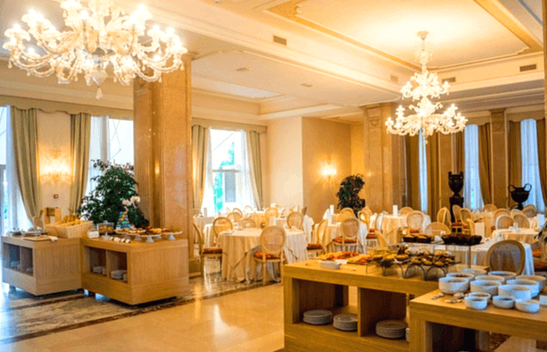 호텔 뷔페 모습이다. 화려한 조명이 천장에 매달렸고, 테이블엔 의자들이 가득하다. 선반엔 다양한 음식들과 그릇들이 있다.