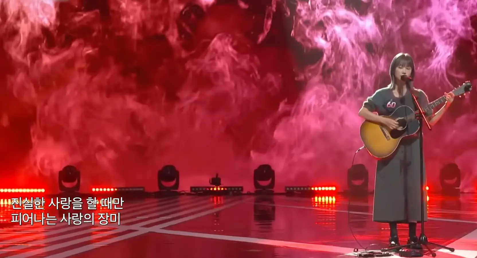 싱어게인 3- 분홍색을 배경으로 키타를 치며 가슴에 60번호표를 달고 단발머리를 늘어뜨린채 노래부르는 60호 전신이미지