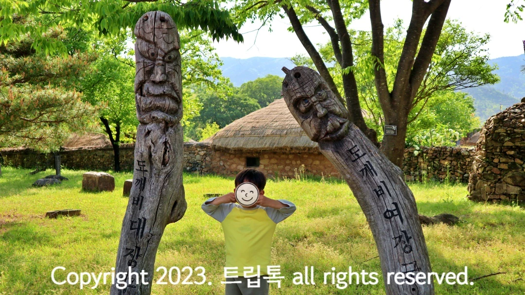 푸른 풀밭에 나무로 만든 장승이 2개 있다. 하나는 휘어서 안쪽을 들여다보는 형상이고 두 장승 사이에 아이가 있다.