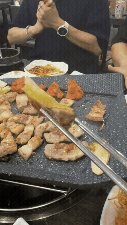 지글지글 익은 제주 오겹살을 백김치와 함께 구워 먹는 장면. 젓가락으로 백김치와 고기를 함께 들어 올린 모습이 먹음직스럽다.