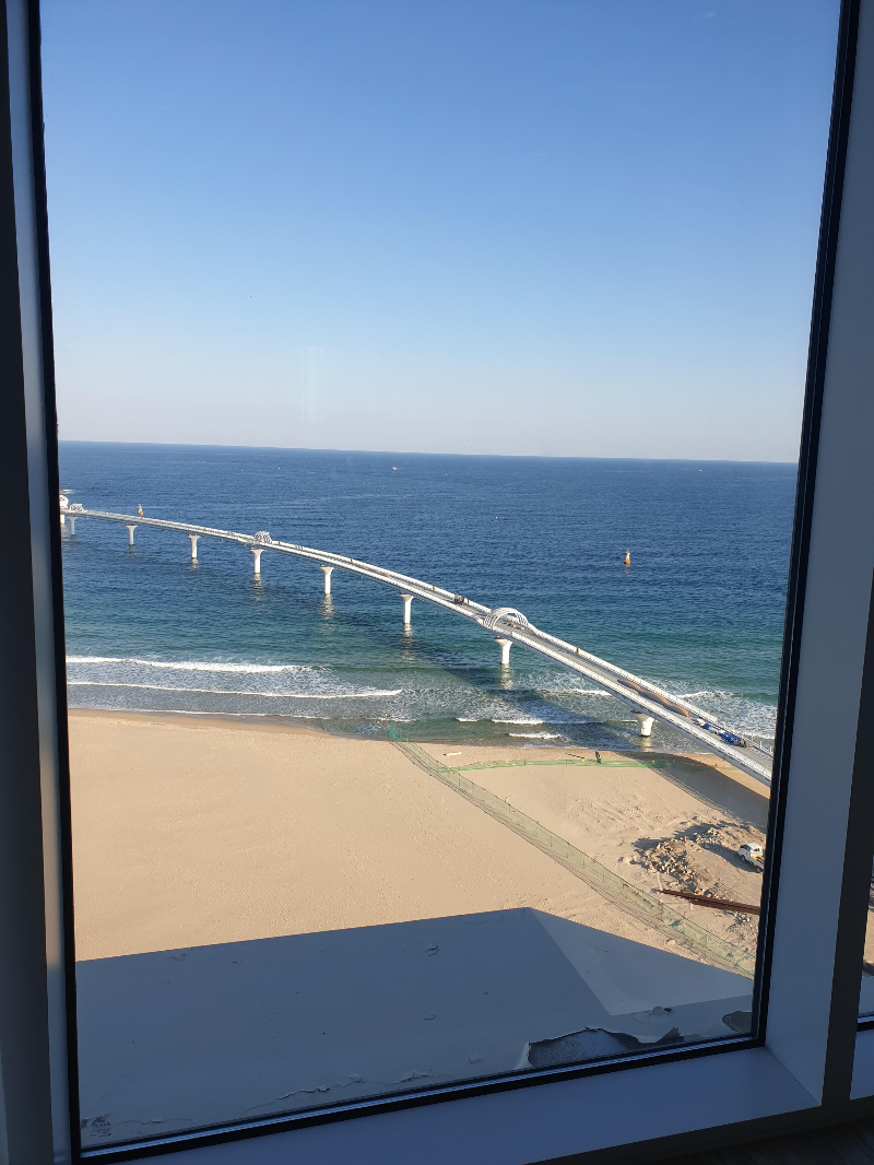 창 밖으로 바다와 해변 그리고 다리가 보인다. 해변 한쪽은 공사 중이다.